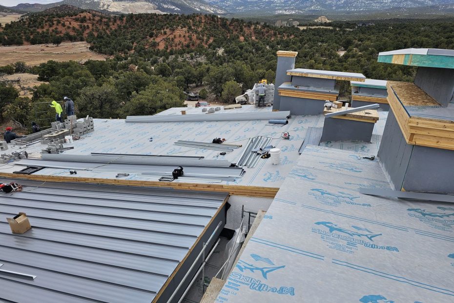 Roof under construction: blue-printed underlayment, staged gray metal panels, workers, mountains and juniper hills beyond