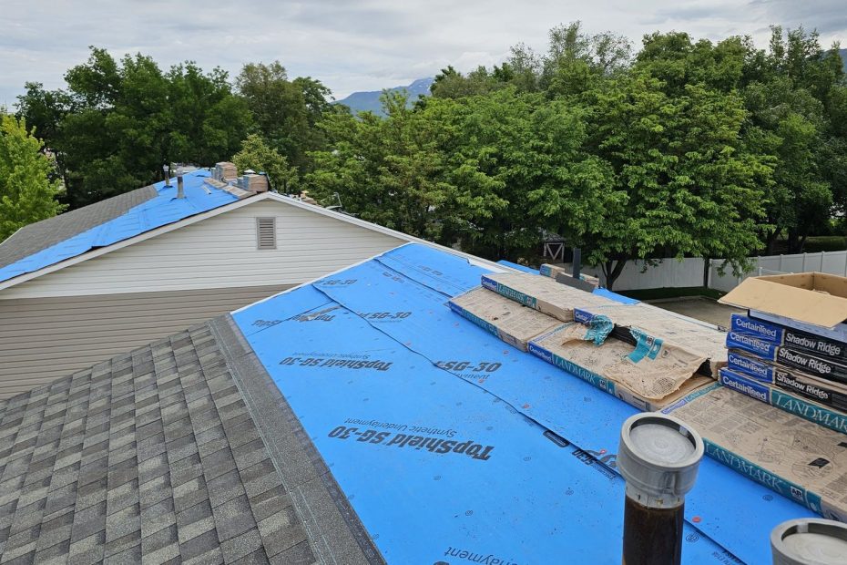 Roof mid-replacement with blue synthetic underlayment, stacked shingle bundles and two metal vent pipes