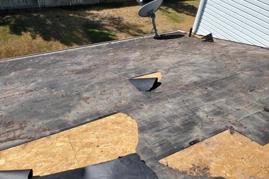 Low-slope roof stripped in patches, dark sheet torn away exposing OSB decking, satellite dish and tear-off shovel
