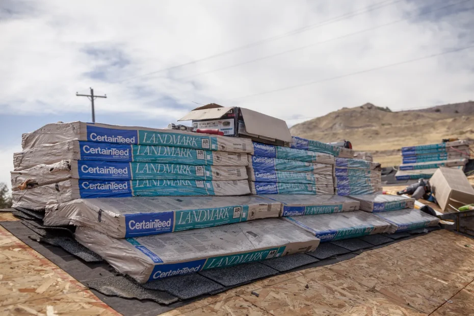 Wrapped CertainTeed Landmark shingle bundles and a nail box stacked on bare plywood roof decking under cloudy sky