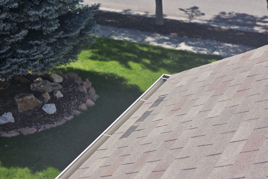 Brown asphalt shingle roof edge with clean white gutter above a green lawn and rock planting bed
