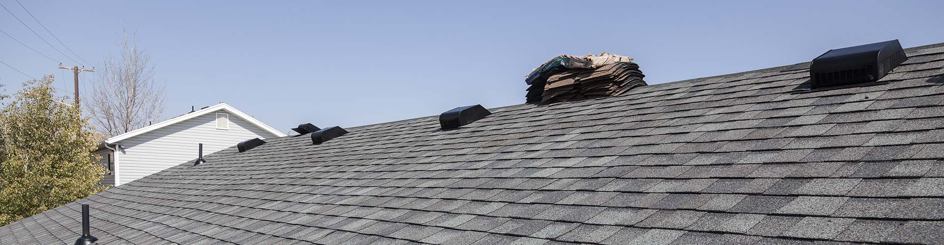 Dark asphalt shingle roof with black box vents and shingle bundles stacked on the ridge under blue sky