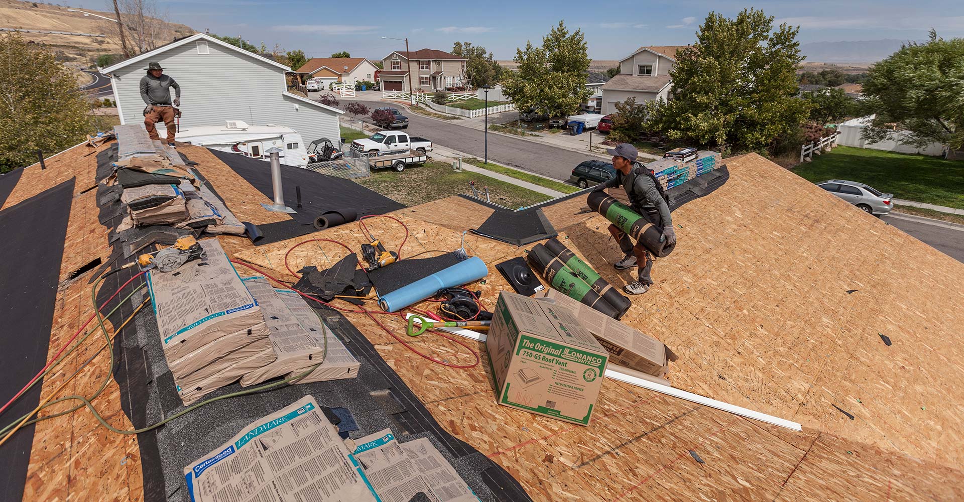 Two roofers on bare plywood deck with underlayment rolls, tools and shingle bundles during a tear-off