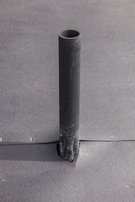 Dark vertical pipe penetrating a gray flat roof membrane, with dark flashing material at its base