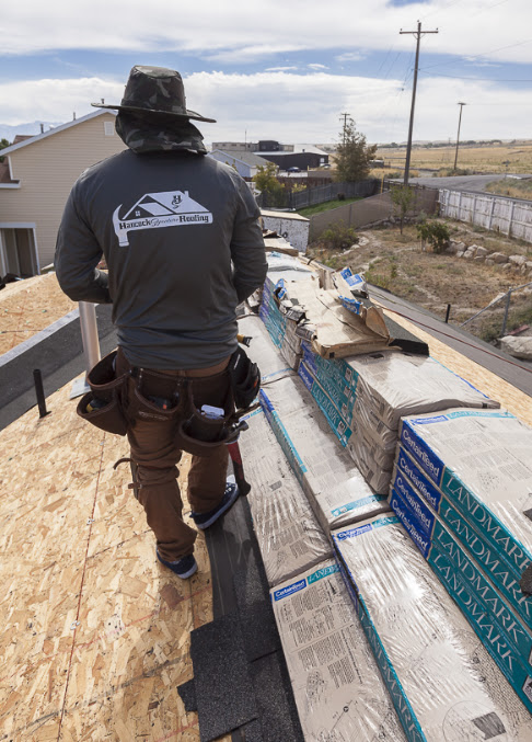 Roofer in branded shirt and tool belt on bare roof deck beside stacked shingle bundles