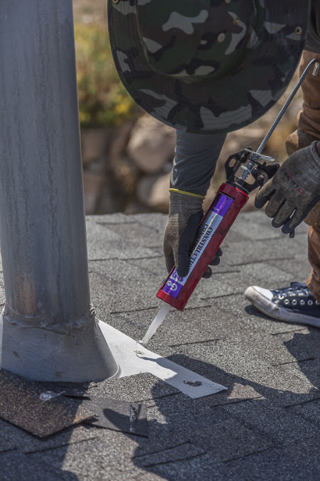 Gloved worker guns sealant around metal flashing at a pipe penetration on an asphalt shingle roof