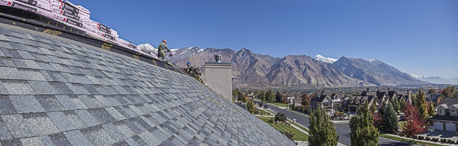 Steep gray shingle roof with two workers and stacked bundles at the ridge, mountains behind