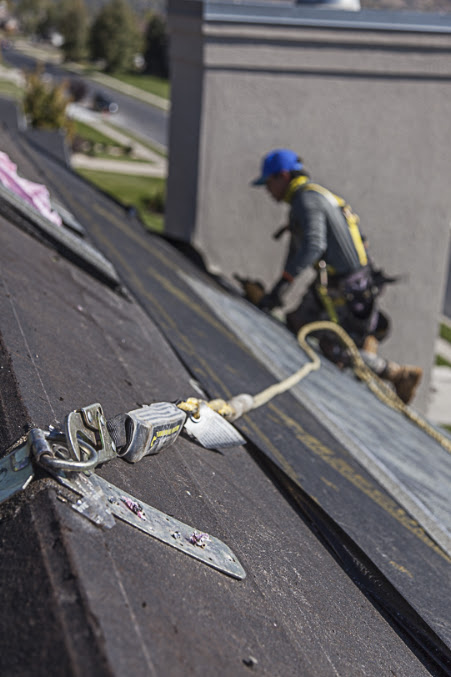 Harnessed roofer on underlayment-covered roof deck with safety lifeline clipped to a foreground anchor plate