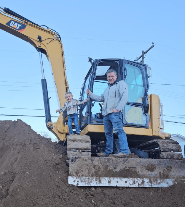 The owner and his young son standing on the tracks of a yellow excavator on a dirt mound