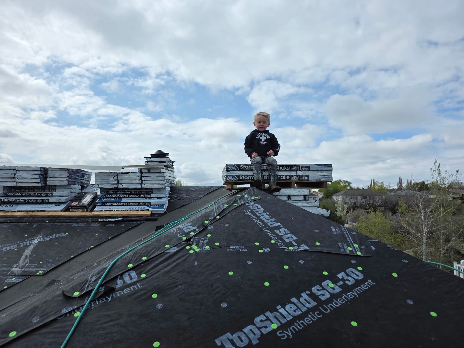 Child sitting on shingle bundle pallet atop a roof covered with black synthetic underlayment