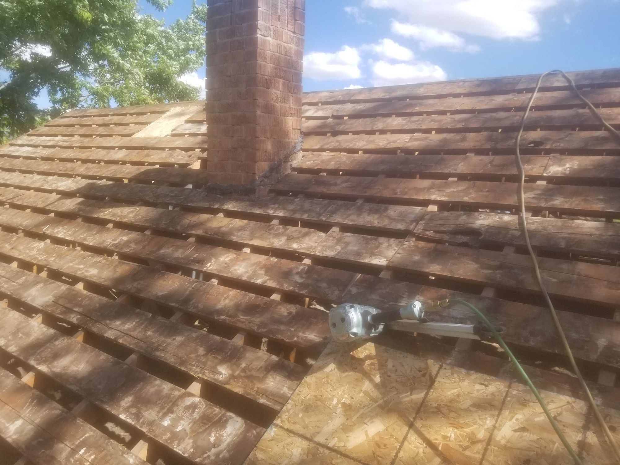 Roof torn off to spaced board decking beside a brick chimney, new OSB sheathing and nail gun nearby