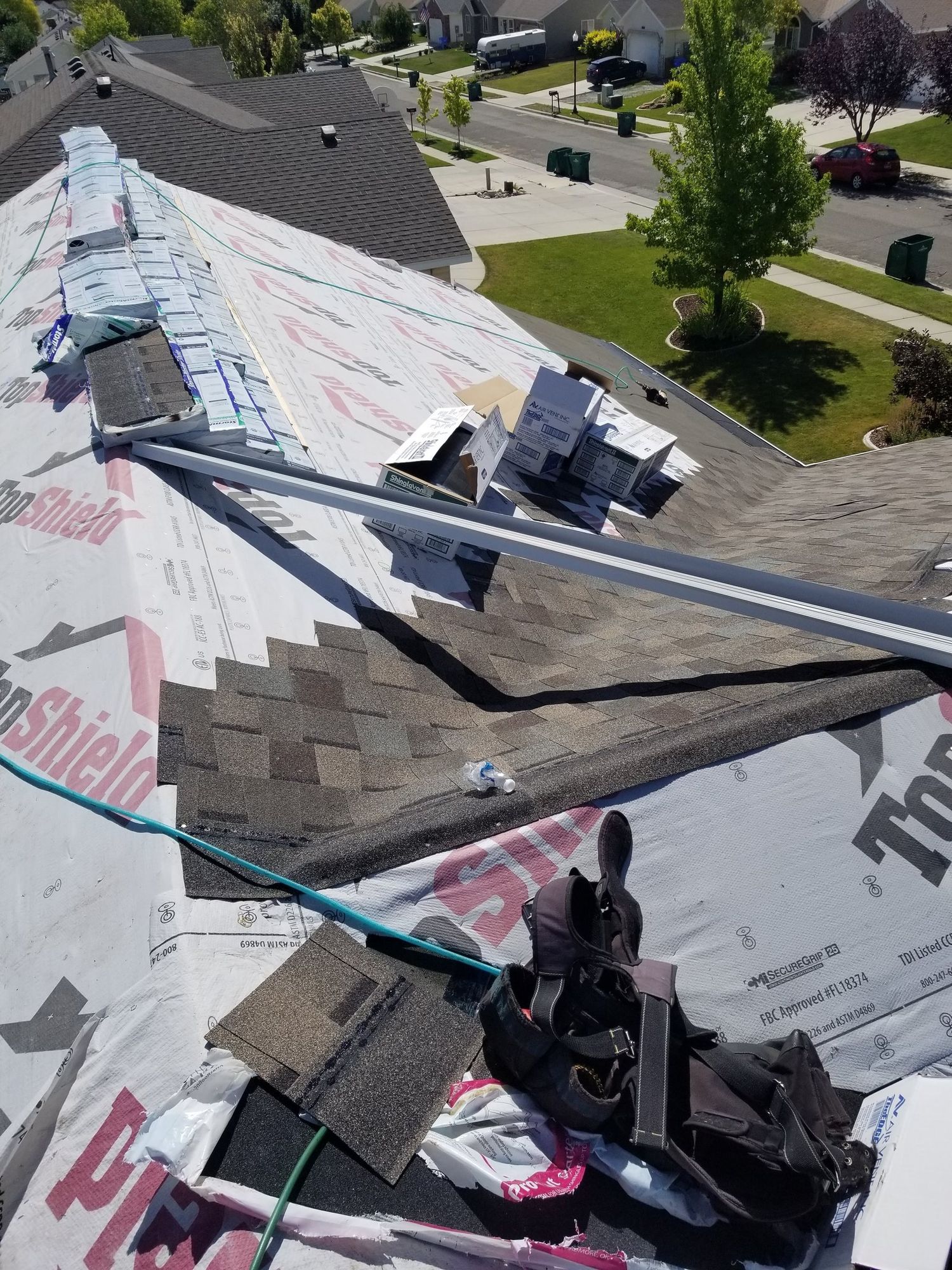 Roof mid-reroof with white underlayment, new asphalt shingle courses, stacked bundles and vent boxes above a suburban street