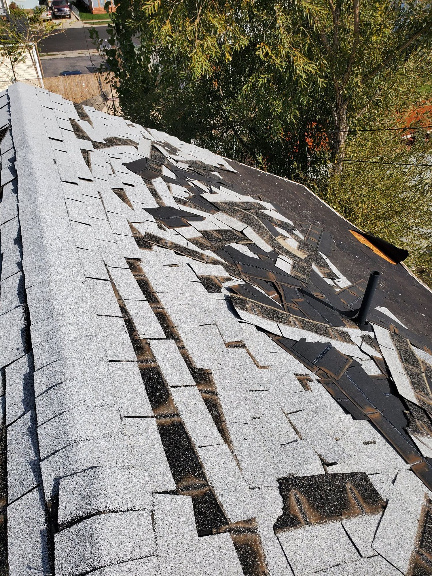 Torn-up gray asphalt shingles along a house ridge exposing black felt and a black vent pipe
