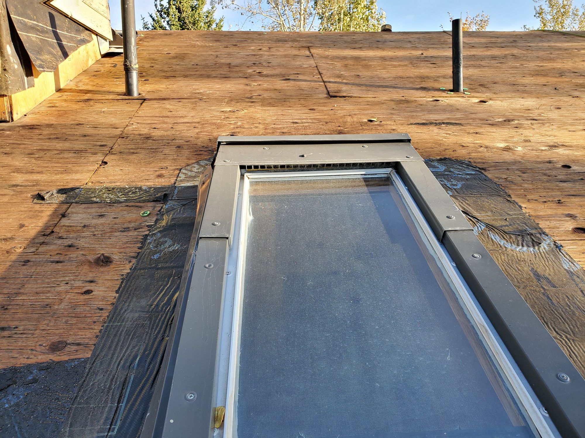 Skylight in bare wood roof decking stripped of shingles, black flashing membrane around it, vent pipes behind, in Kearns, UT