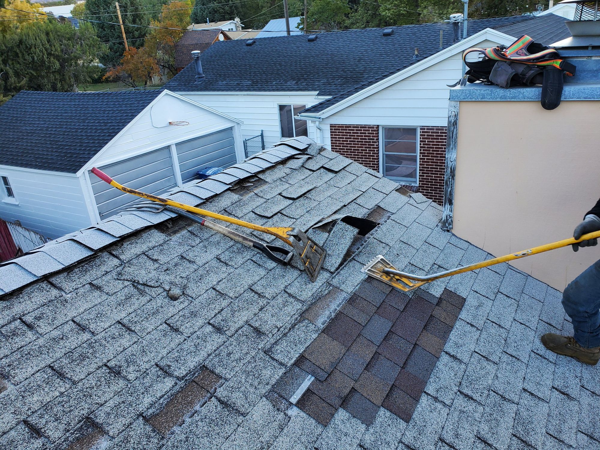 Roofer using tear-off shovel to strip gray asphalt shingles near a ridge, second shovel lying on roof