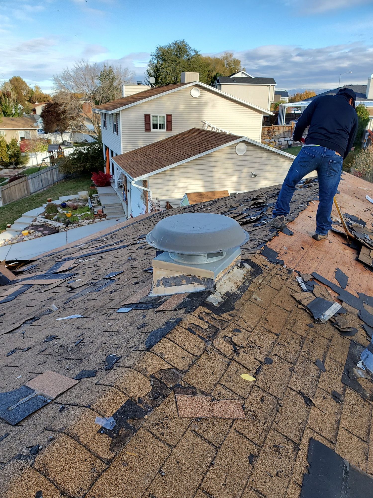 Roofer stripping old asphalt shingles around a domed vent, bare plywood decking exposed near the ridge, in Kearns, UT