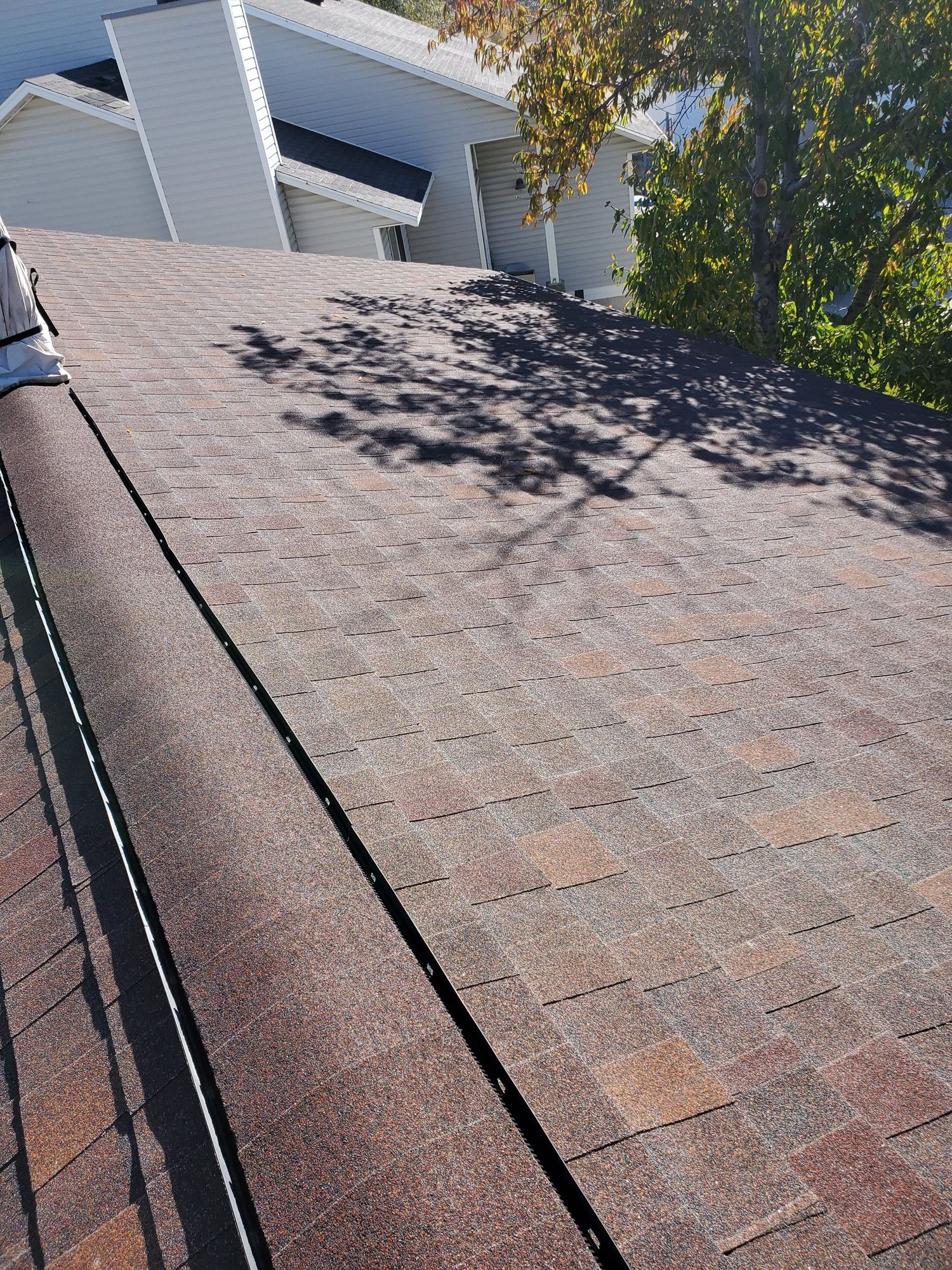 Brown asphalt shingle roof plane with dark metal valley, dappled tree shadows, white-sided house beyond, in West Haven, UT