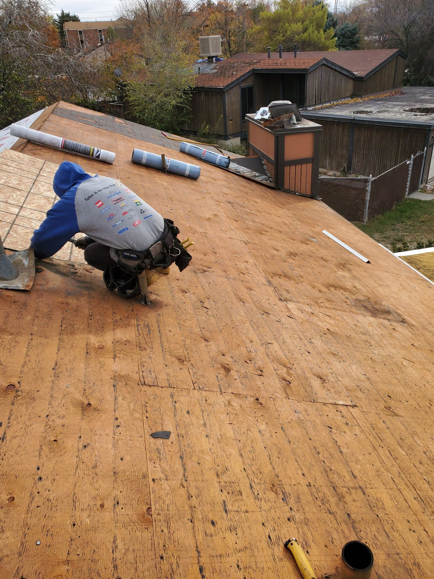 Roofer kneeling on bare plywood roof deck beside rolls of underlayment during a tear-off