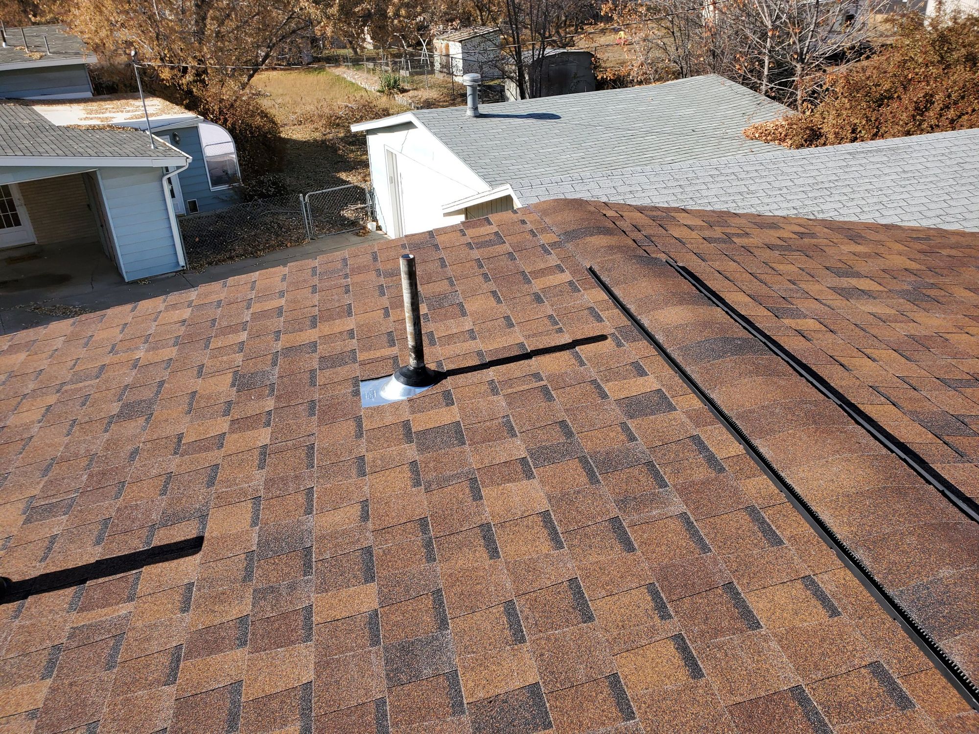 New brown asphalt shingle roof with metal pipe flashing and uncapped black ridge vent along the ridge