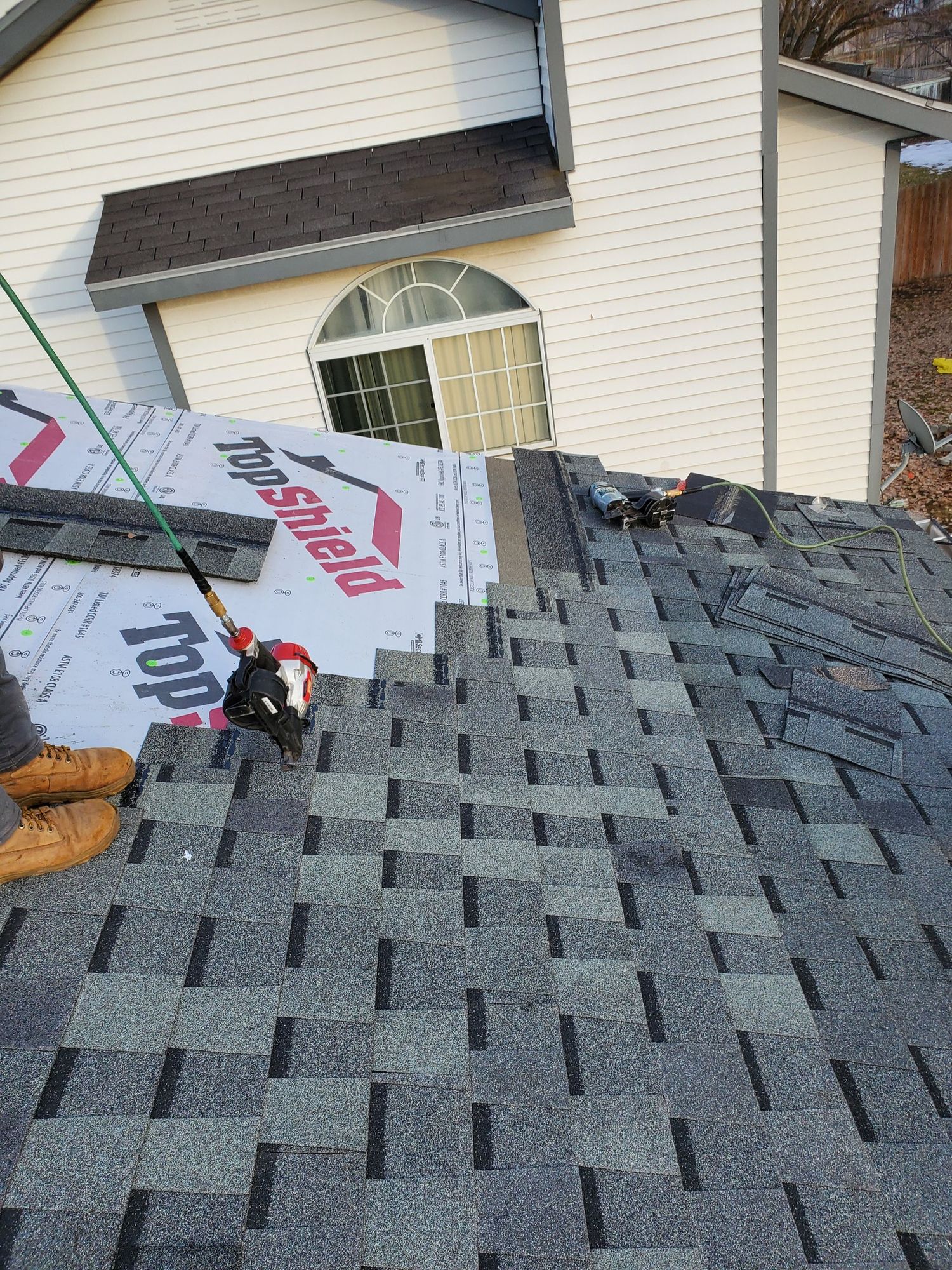 Steep roof mid-shingling: gray shingles, white Top Shield underlayment, two nail guns, worker's boots at edge