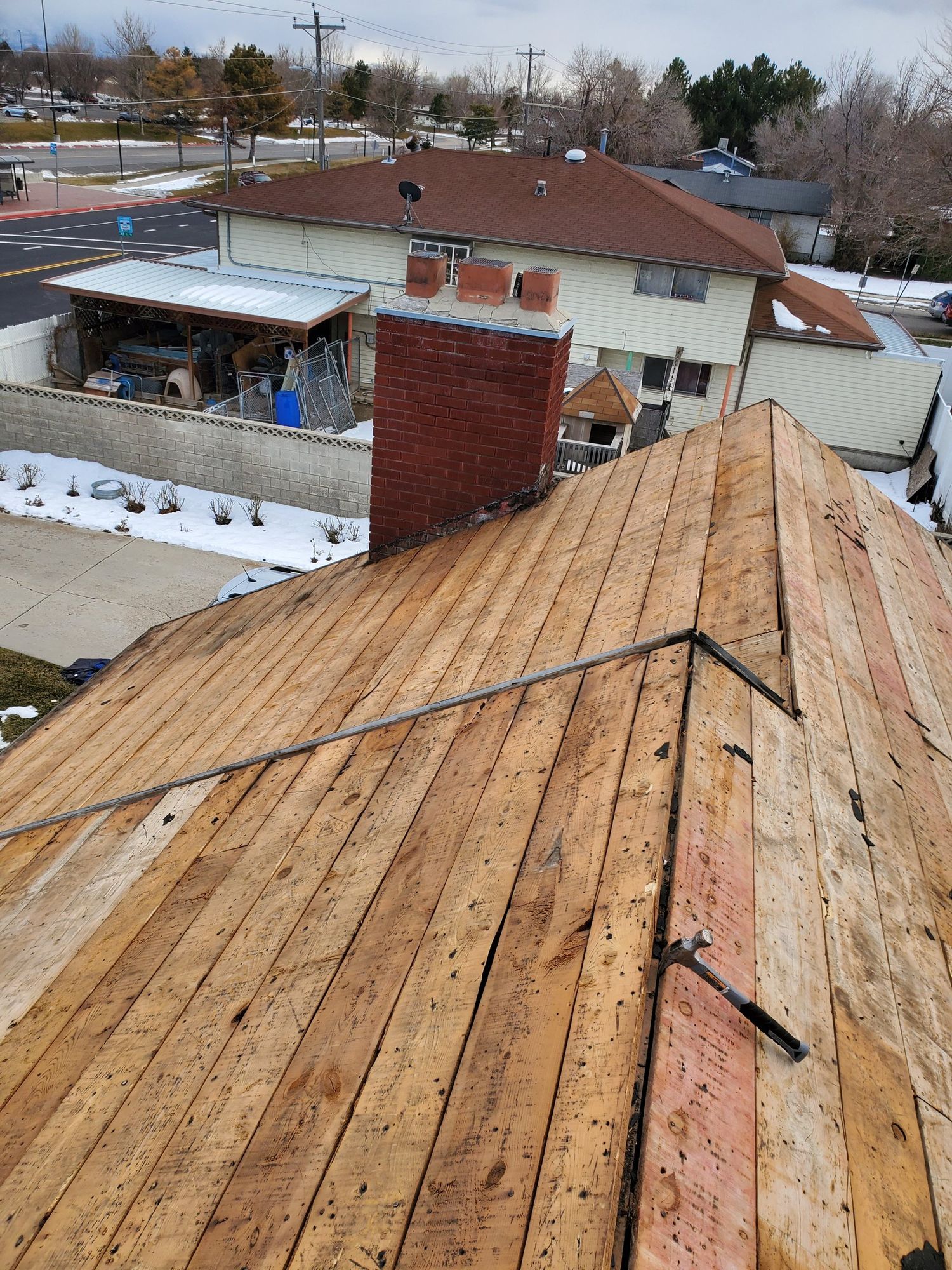 Roof torn off to bare wood plank decking, hammer on slope, brick chimney and snowy neighborhood behind