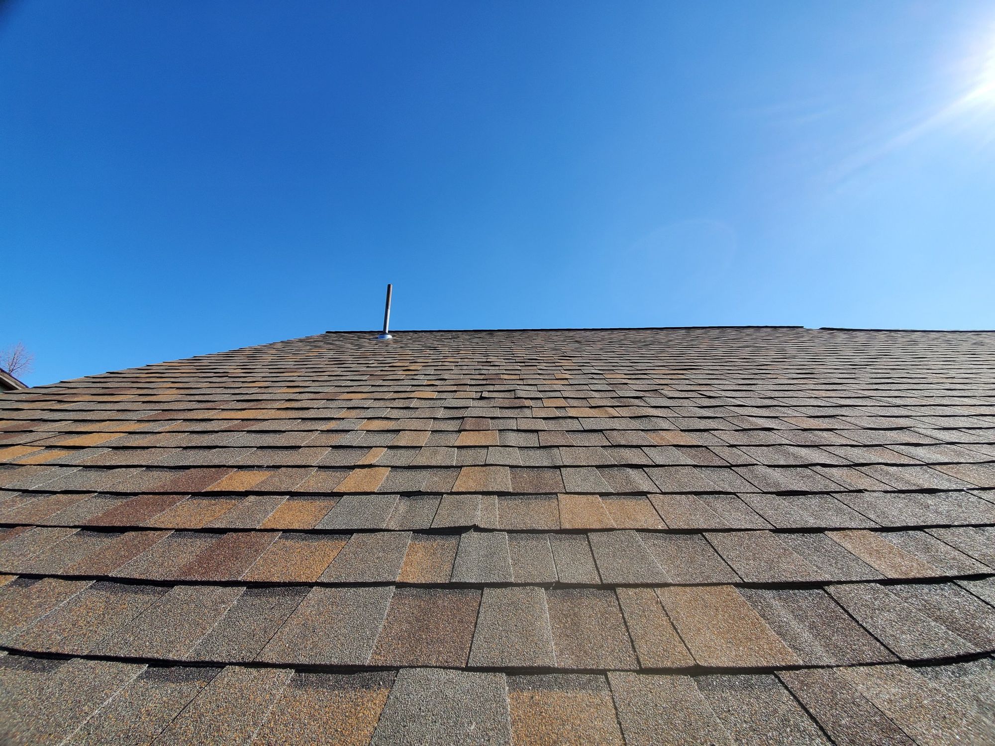 Brown asphalt shingle roof slope rising to a ridge with a metal flue pipe, blue sky above