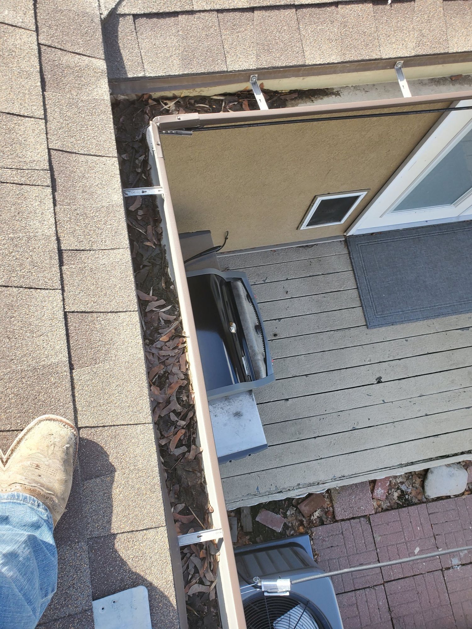 Gutter along shingle roof eave clogged with dead leaves, deck, grill and AC unit below