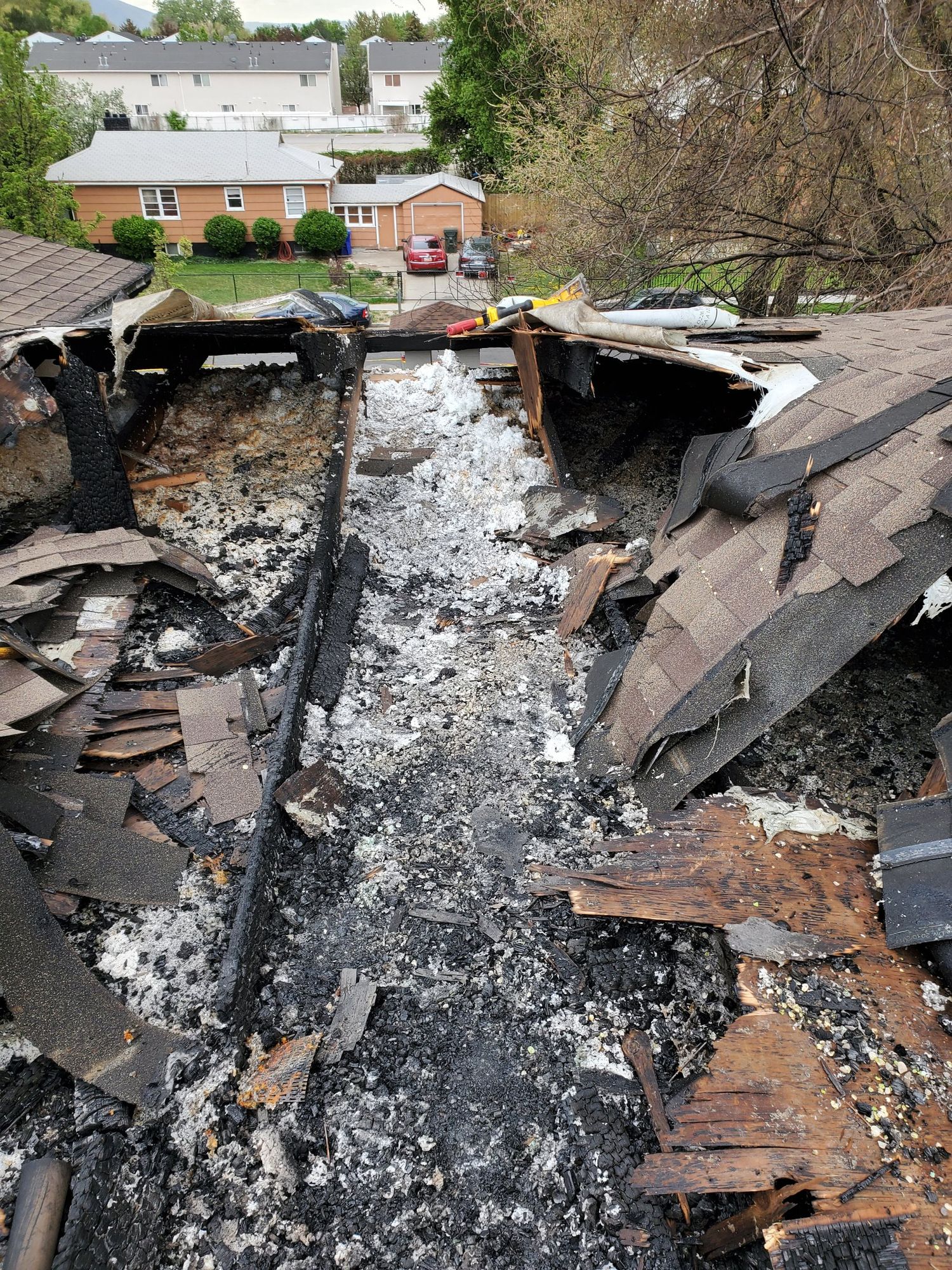 Fire-destroyed roof section with charred rafters, ash and collapsed brown asphalt shingles, houses visible beyond, in West Jordan, UT