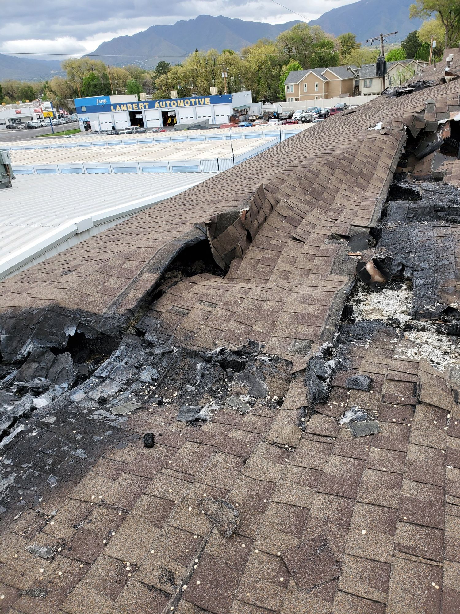 Fire-damaged brown asphalt shingle roof with burned holes and charred decking near the ridge, commercial buildings beyond