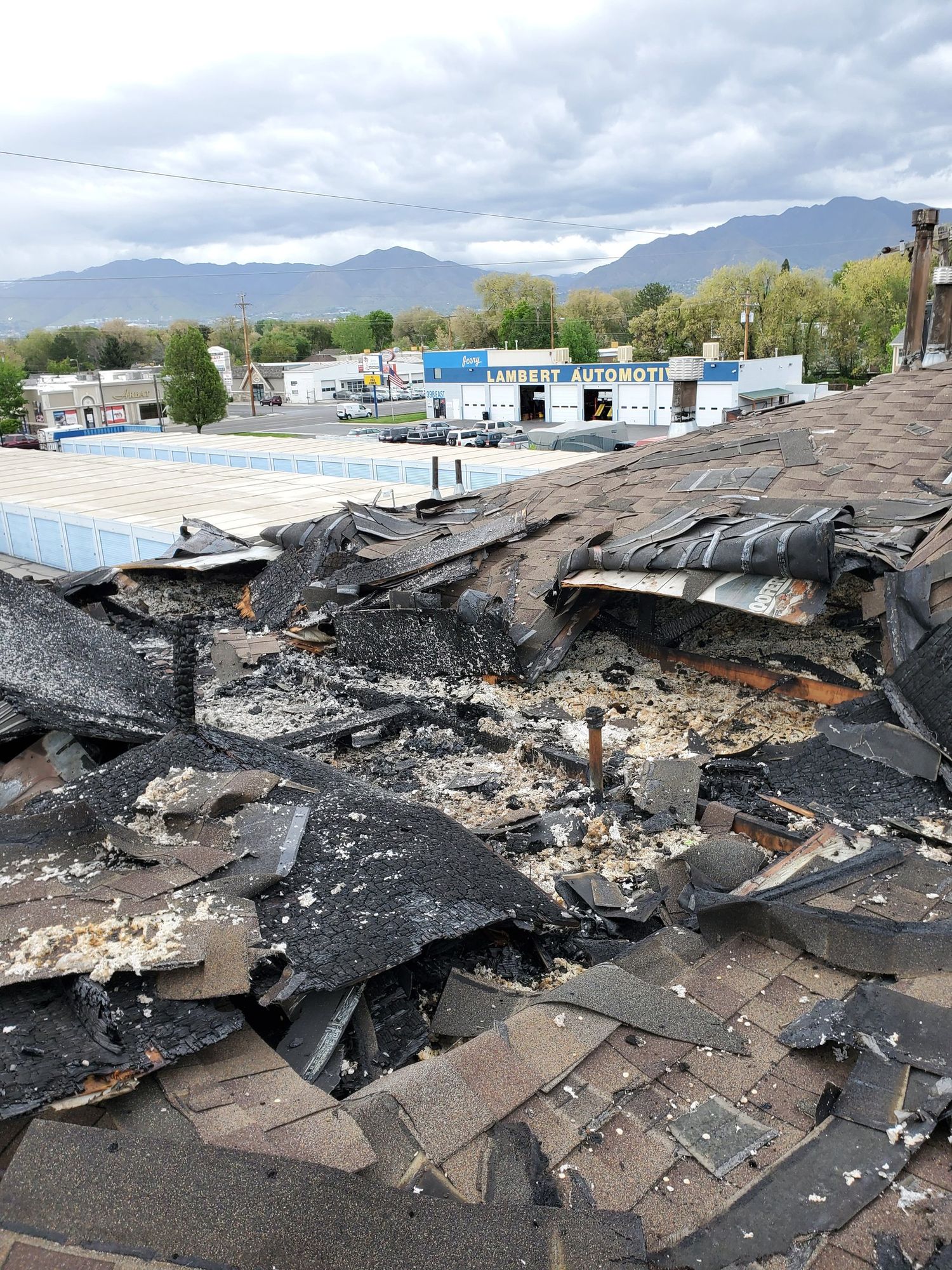 Burned-through asphalt shingle roof with charred debris and insulation, auto shop and mountains beyond, in Farmington, UT