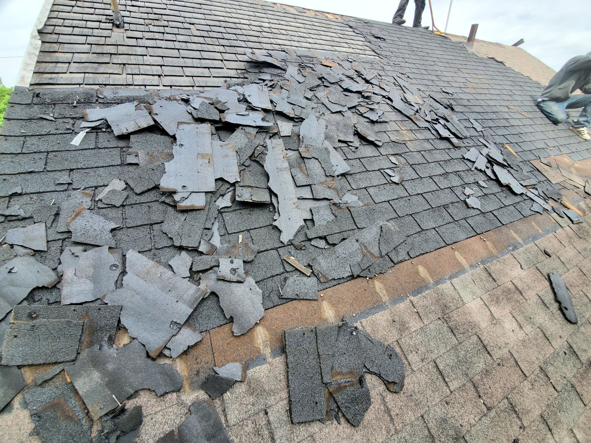 Stripped asphalt shingle debris covering a steep roof slope mid tear-off, workers near the ridge above, in Millcreek, UT