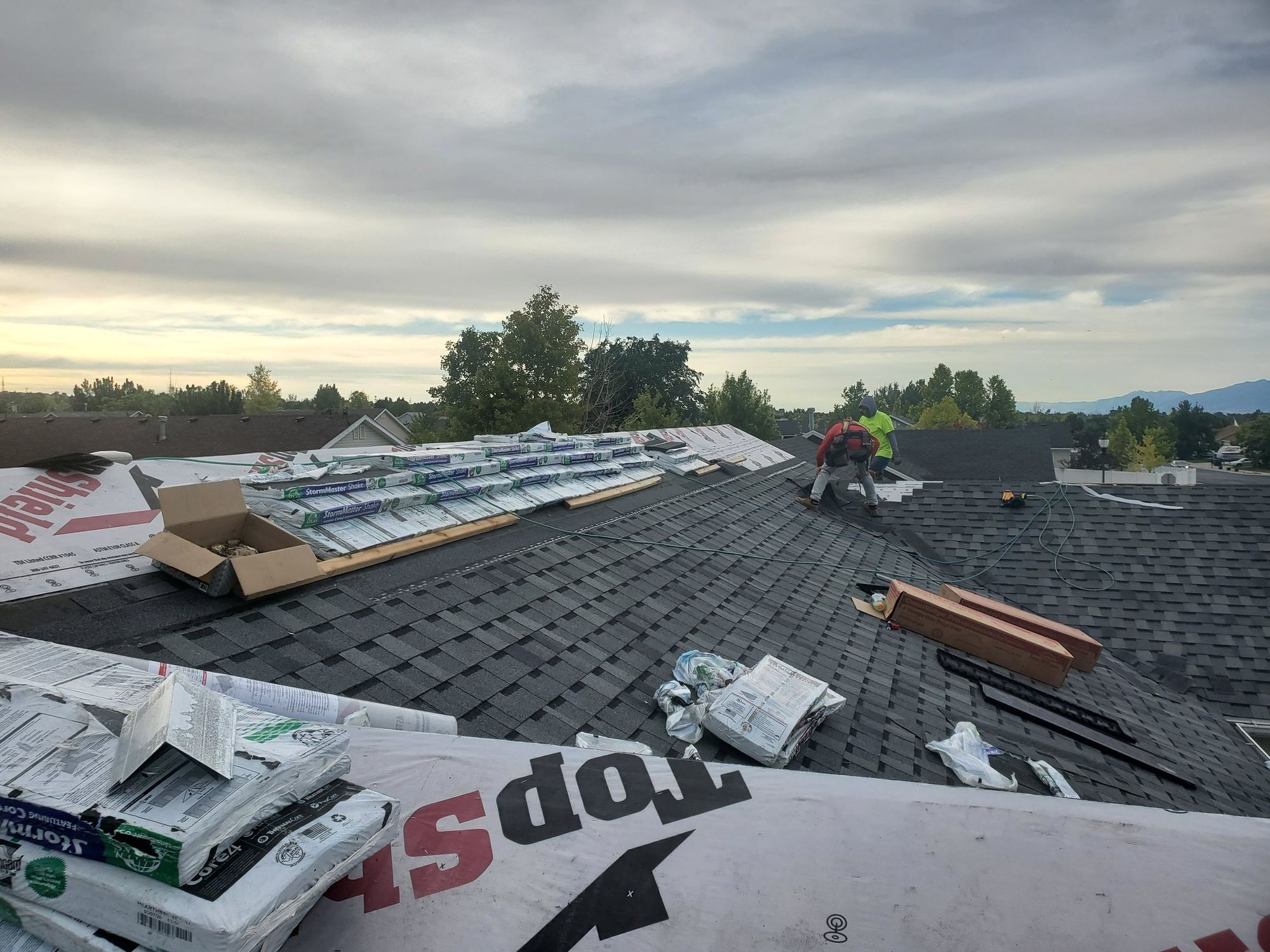 Workers on a residential asphalt shingle roof with staged shingle bundles and underlayment wrappers around them
