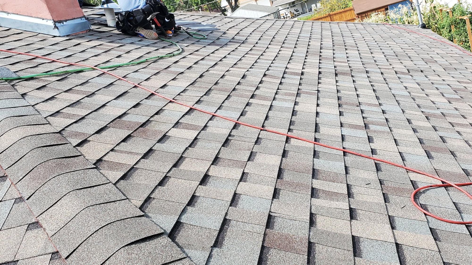 New asphalt shingle roof plane with air hoses; worker's legs and tool belt visible near chimney at ridge