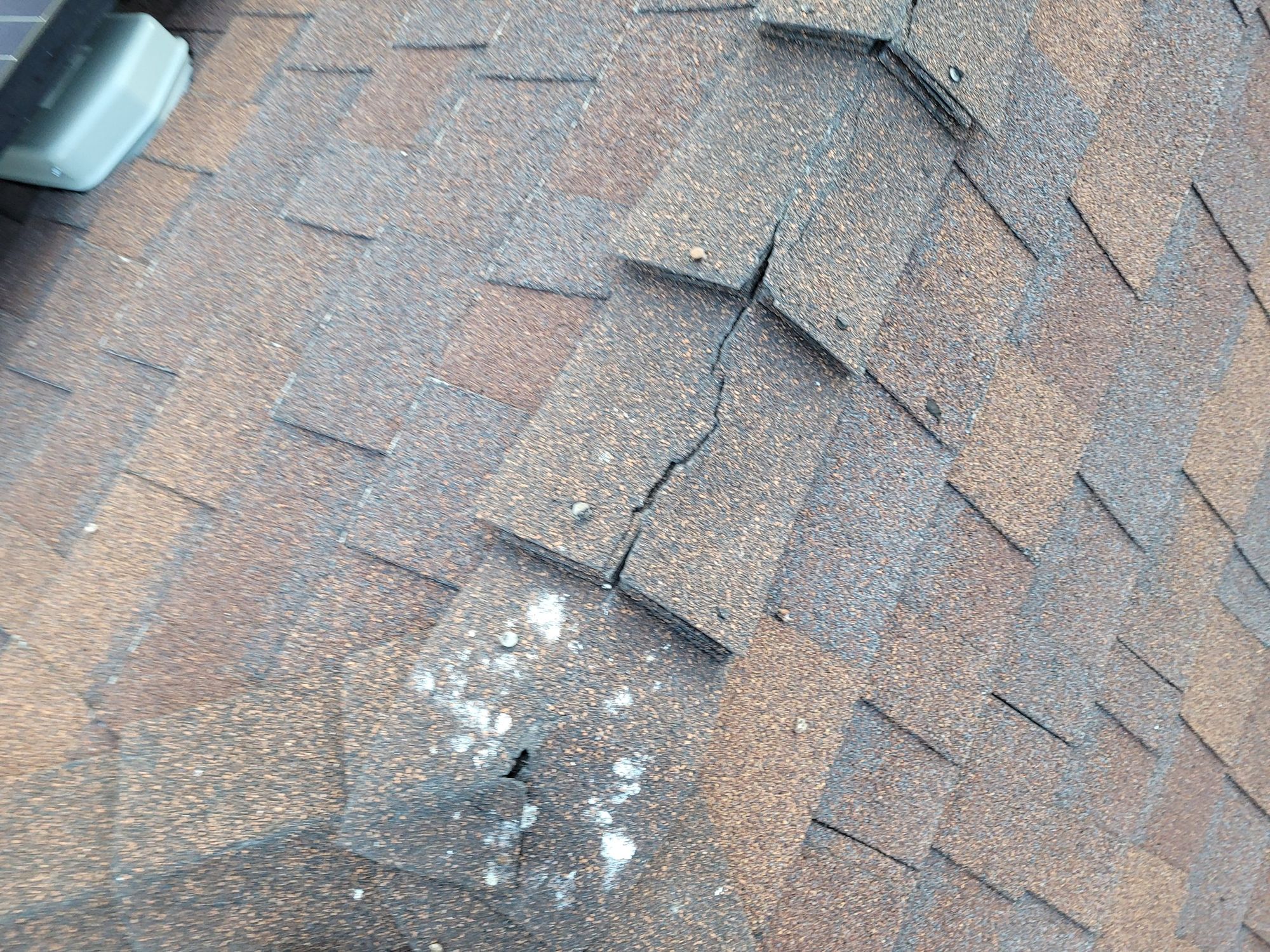 Weathered brown asphalt shingles with a long crack, lifted tabs, exposed nail heads and white staining
