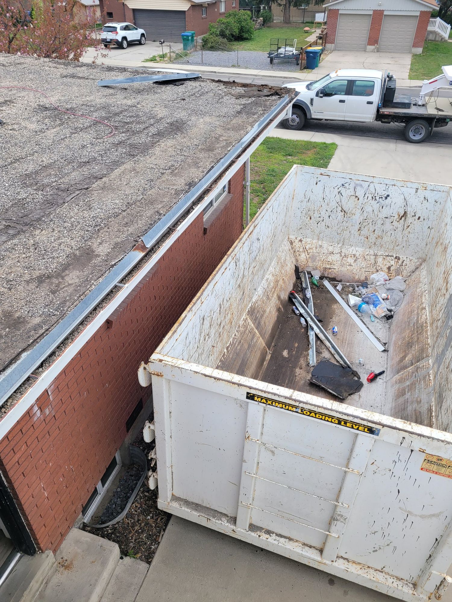 Gravel low-slope roof edge with loose metal trim above a white roll-off dumpster in a driveway