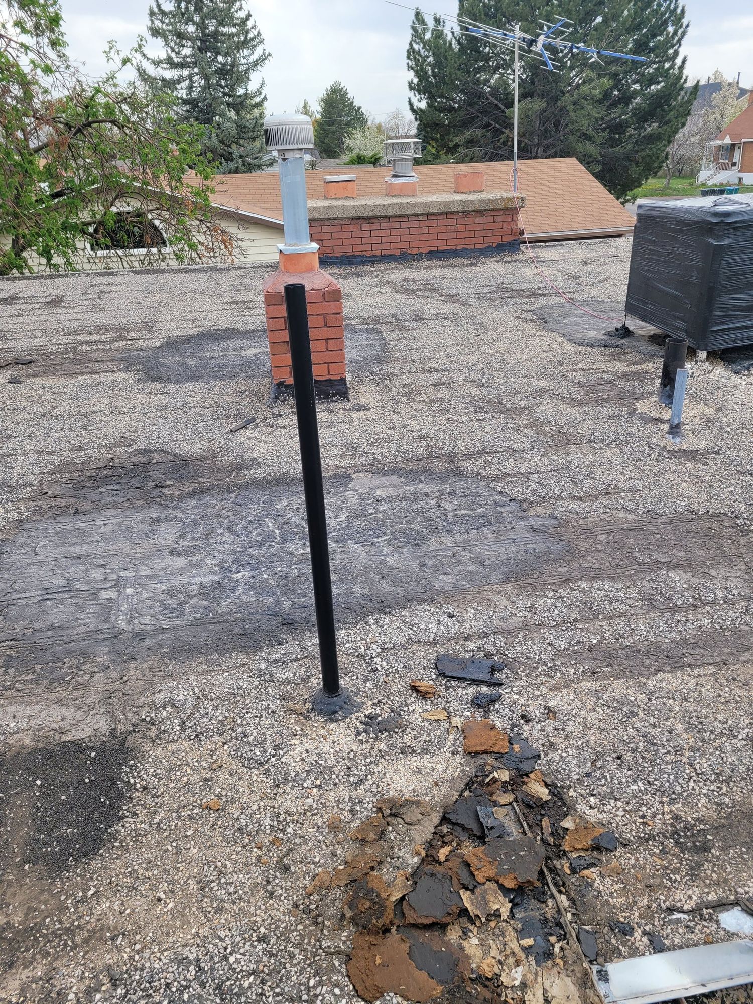 Worn flat gravel roof with bare membrane patches, torn felt debris, black vent pipe and brick chimney