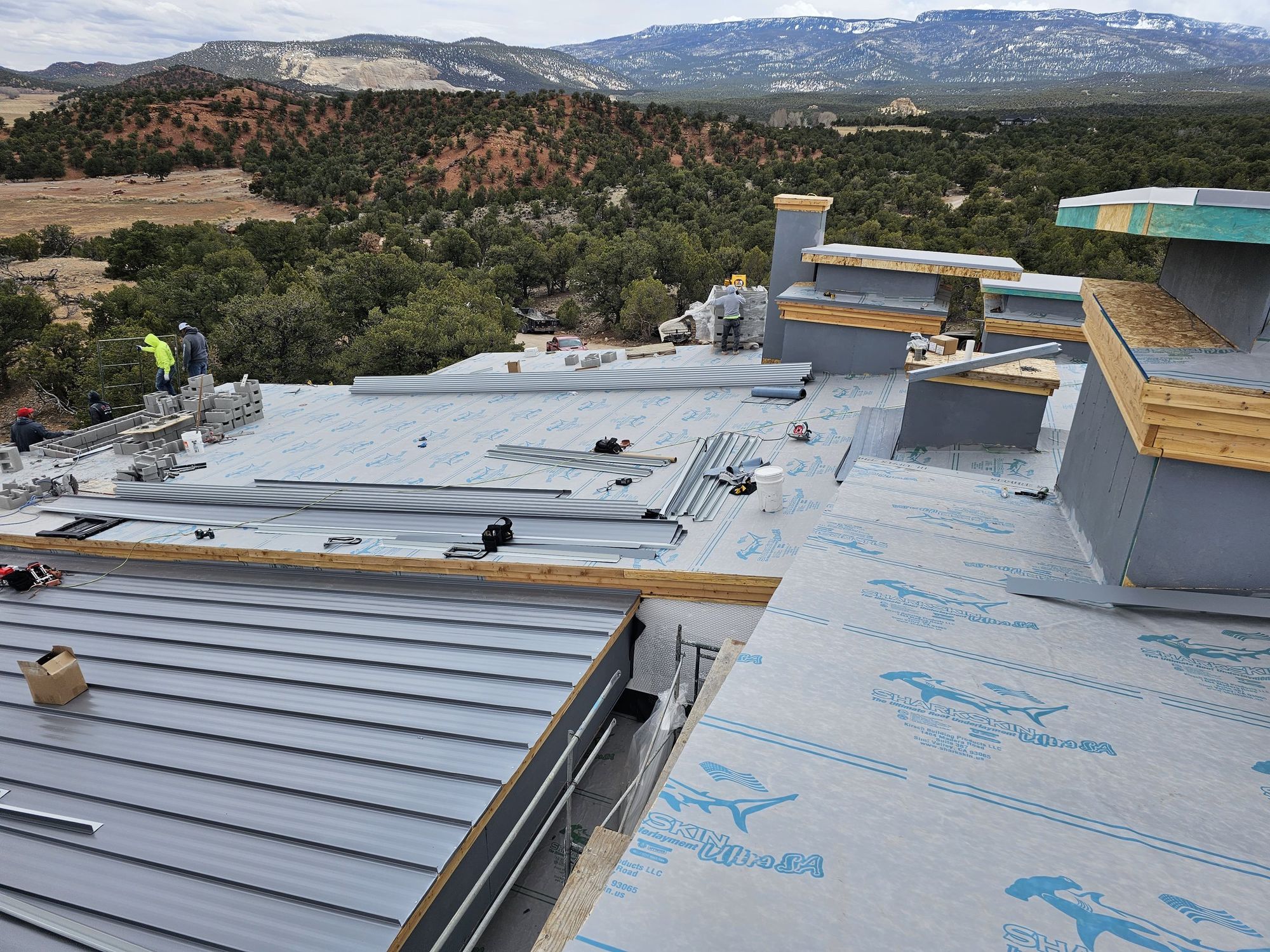 Roof under construction: blue-printed underlayment, staged gray metal panels, workers, mountains and juniper hills beyond