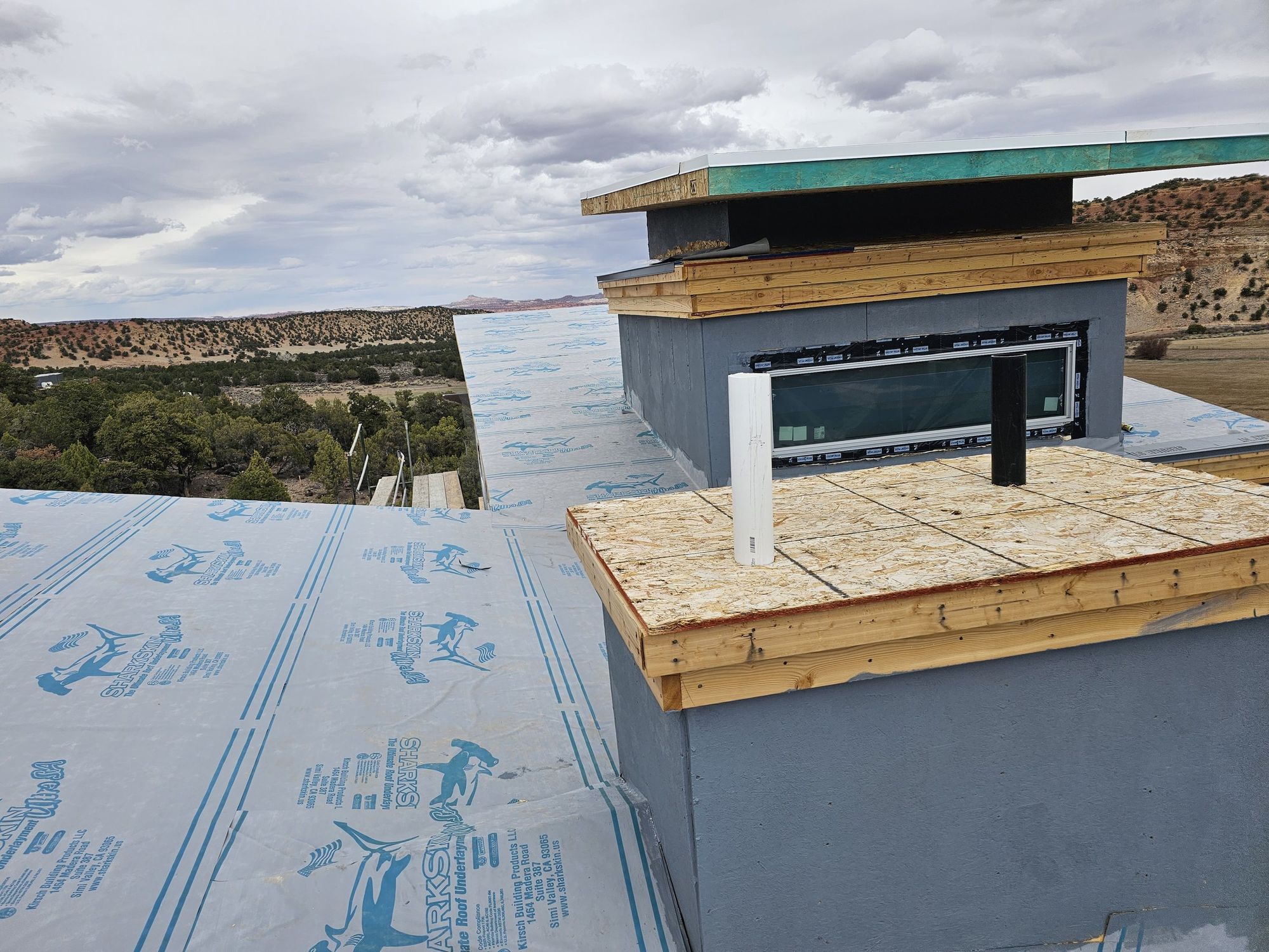 In-progress roof: blue synthetic underlayment, bare OSB decking, pipe penetrations, new wood trim, desert valley beyond
