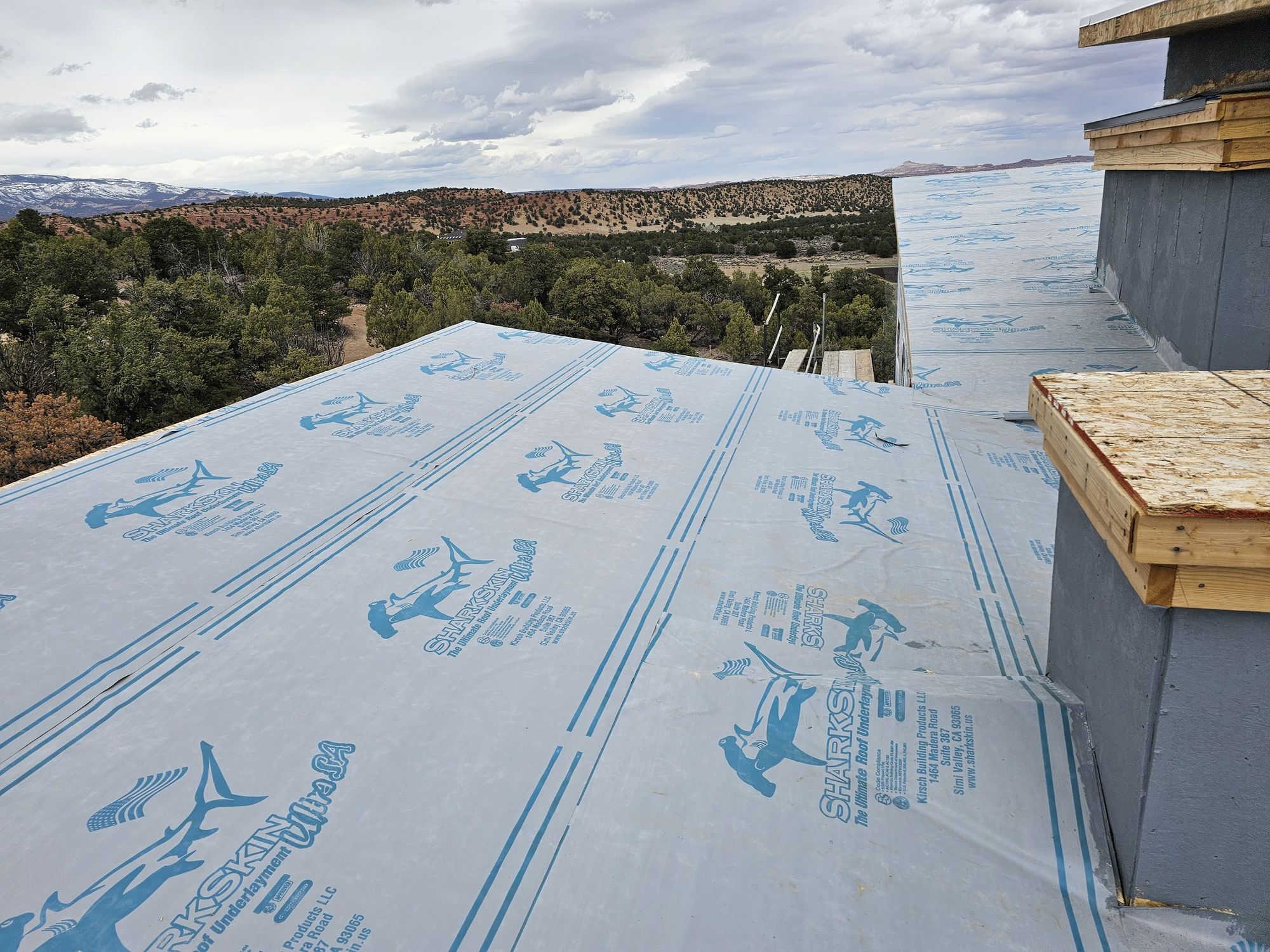 Roof deck covered with blue-printed Sharkskin underlayment, gray parapet walls, wooded hills and cloudy sky beyond