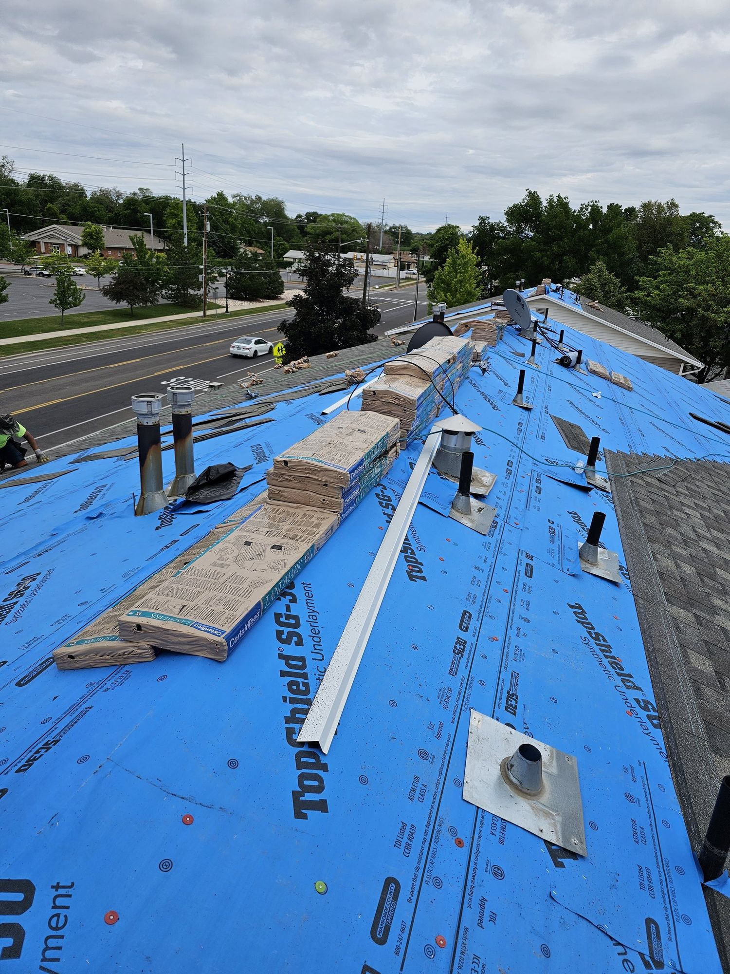 Roof covered in blue synthetic underlayment with shingle bundles stacked along the ridge near metal vent pipes