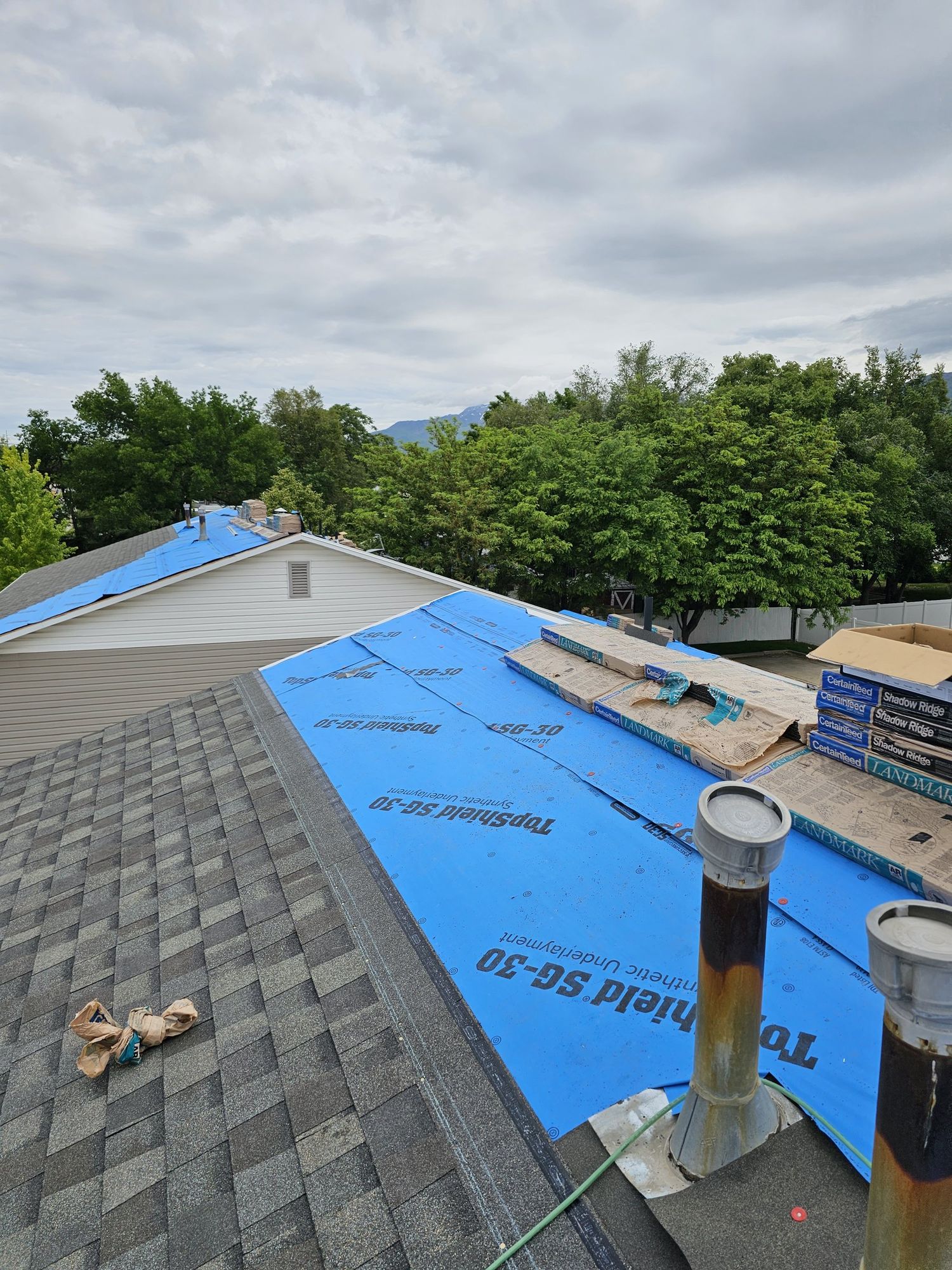 Roof mid-replacement with blue synthetic underlayment, stacked shingle bundles and two metal vent pipes