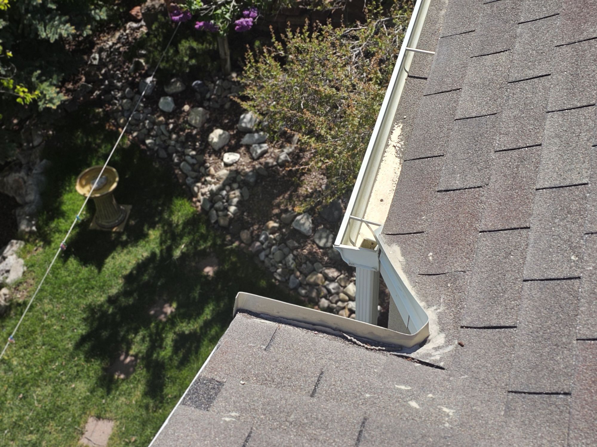 Looking down on a cream gutter corner and downspout at an asphalt shingle roof eave