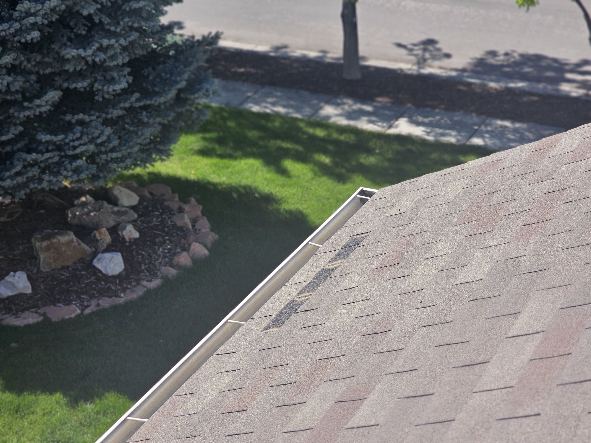 Brown asphalt shingle roof edge with clean white gutter above a green lawn and rock planting bed