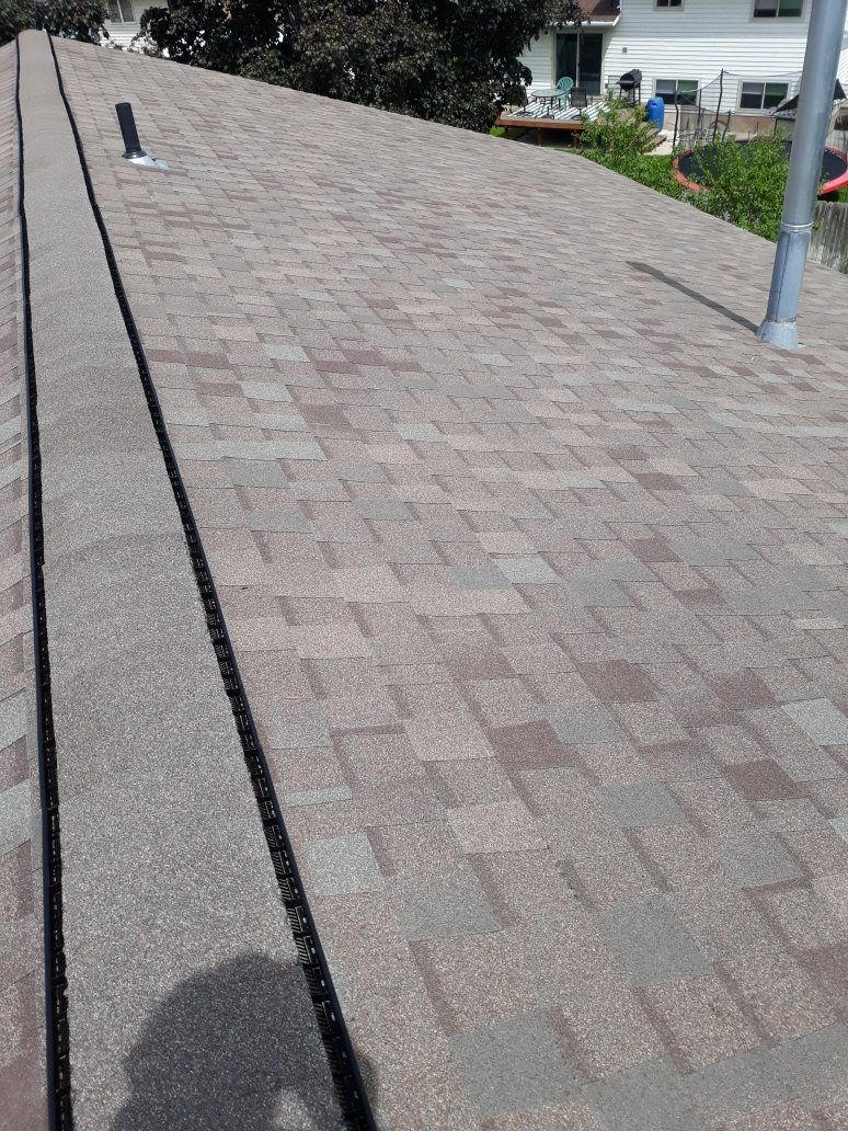 Asphalt shingle roof plane with exposed black ridge vent, vent pipe and metal flue, houses behind