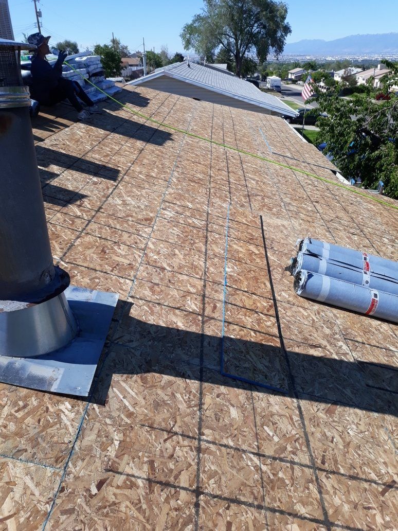 Bare OSB roof deck with chalk lines, underlayment rolls, flue pipe flashing and a seated worker by shingle bundles