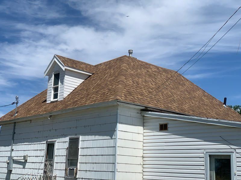 White-sided house with tan asphalt shingle hip roof, small dormer window and metal ridge vent against blue sky