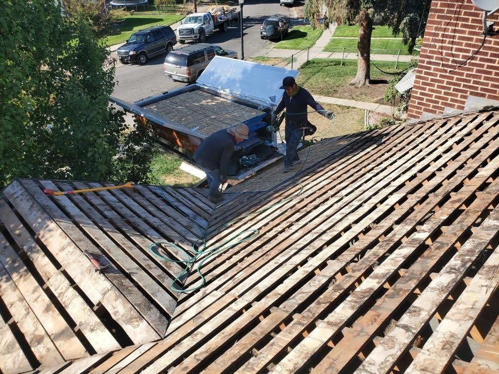 Workers on a roof stripped to bare wood slats, debris trailer parked at the eave below, in South Jordan, UT