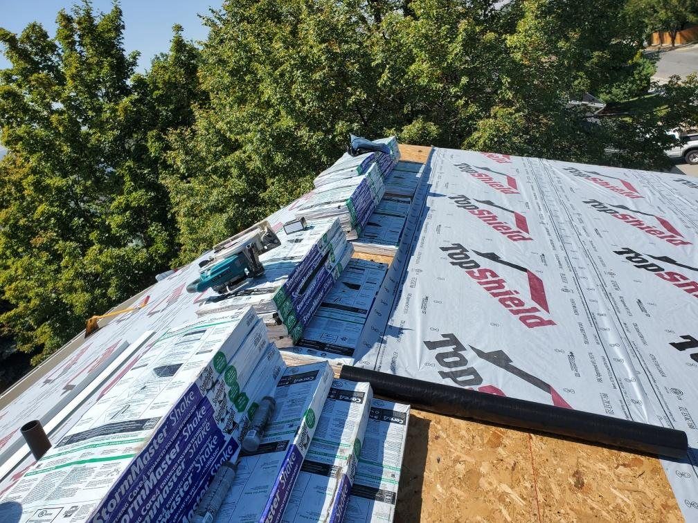 Roof deck covered in white synthetic underlayment with stacked wrapped shingle bundles and tools along the ridge