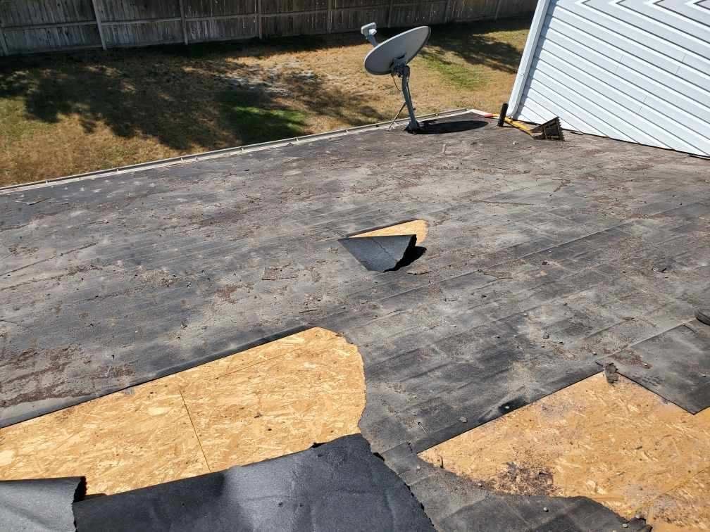 Low-slope roof stripped in patches, dark sheet torn away exposing OSB decking, satellite dish and tear-off shovel