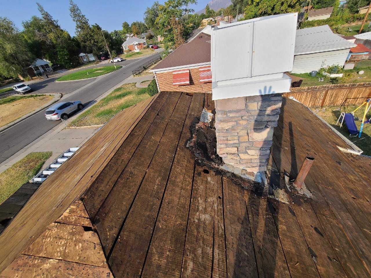 Stripped roof deck of bare boards beside a stone chimney with rotted wood and failed flashing